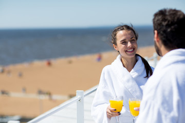 Cherful young wife with glass of juice looking at husband while talking to him at spa resort on...