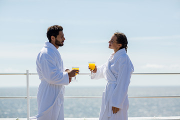 Young couple in bathrobes having talk by drink while standing by waterside on sunny day