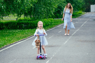 little and sweet blonde girl in a cute gray dress riding a scooter together with beautiful mother...