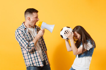 Angry fun expessive couple, woman man football fans screaming, cheer up support team with soccer ball, megaphone isolated on yellow background. Sport family leisure quarrel problem lifestyle concept.