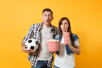 Young fun couple, woman man, football fans holding soccer ball, bucket of popcorn, plastic glass of cola, cheer up support team, isolated on yellow background. Sport family leisure lifestyle concept.