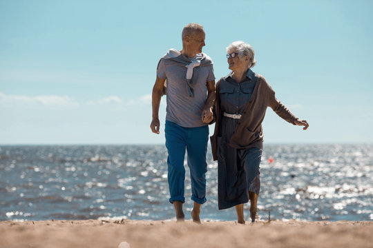 Couple of senior man and woman having talk while moving along sandy beach at summer resort