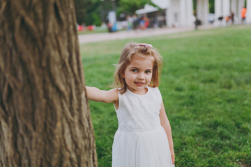 Portrait of smiling little cute child baby girl in light dress stand and lean on tree on green grass in park. Mother, little kid daughter. Mother's Day, love family, parenthood, childhood concept.