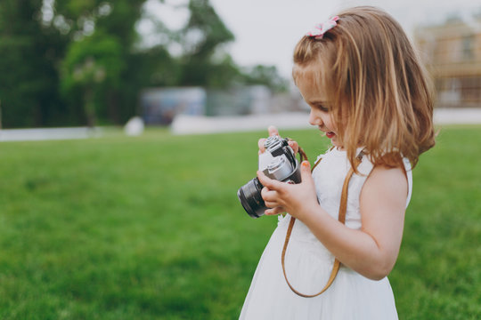 Little pretty child baby girl in light dress holding retro vintage photo camera on green grass in city park. Mother, little kid daughter. Mother's Day, love family, parenthood, childhood concept.
