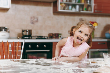 Little funny kid girl in pink dress playing, smearing hands and face with flour in light kitchen at table. Child daughter cooking food, cookies in weekend morning at home. Happy childhood concept.