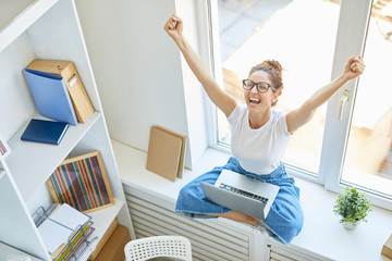 Excited and happy young woman with laptop sitting on window sill with her hands raised and...
