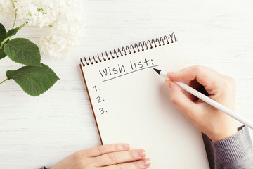 Woman's hand writing Wish list in notebook on white wooden table. Working place and planning...