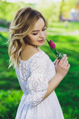 Young beautiful blonde girl in white dress with a flower tulip on a background of green grass