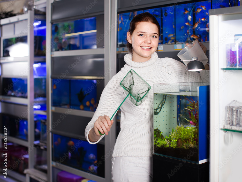 Wall mural Girl holding aquarian net and water container next to aquarium with colorful fish in aquarium shop