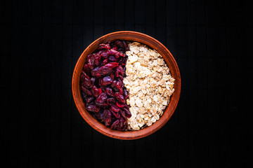 Raw oatmeal with dried cranberries in a round clay bowl on a dark wooden background. Healthy fitness vegetarian breakfast granola meal with cranberries. Top view