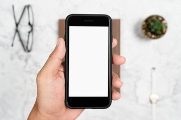 Hand holding smartphone white screen for mockup design or app display with blurred eyeglasses and flower on desk backgrounds