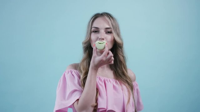 Good-looking young woman in light pink dress bites two macaroons, actively chewing, covering her mouth via macaroon on light blue colored background. Having fun, delicious, being a sweet tooth. Female