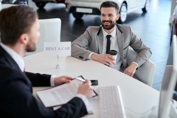 Smiling bearded businessman with coffee sitting at auto salon and communicating with salesman. He wants to rent a car