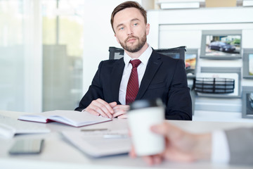 Serious young salesman sitting at desk with notebook and listening to his client