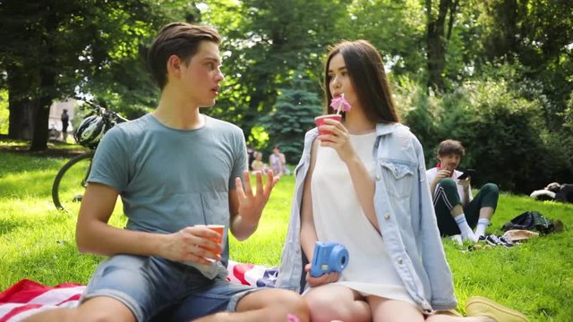 Good-looking young man telling story to beauty. Attractive Caucasian girl in romantic dress listening attentively to boyfriend, holding camera in hand, drinking juice with straw.