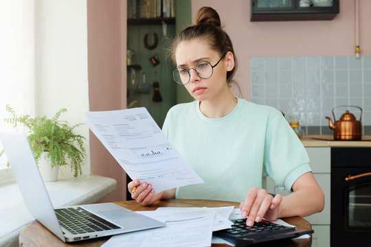 Closeup picture of young attractive girl with glasses and no makeup spending afternoon at home analyzing data in important financial documents looking concentrated, serious and uneasy having problems