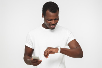 Closeup shot of African American man pictured isolated on grey background listening to music through wireless earphones, using smartphone and looking at digital smart wristwatch, fan of gadgets