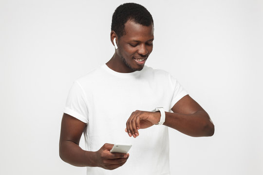 Horizontal photo of young African American man isolated on gray background dressed in blank white T-shirt using simultaneously phone and digital wristwatch as if trying to adjust its settings