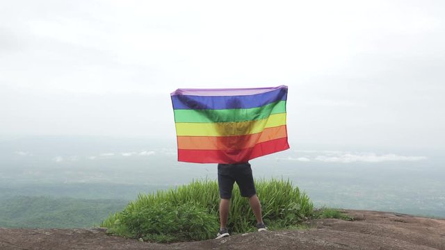 man waving rainbow flag LGBTI flag in hard wind on mountain top