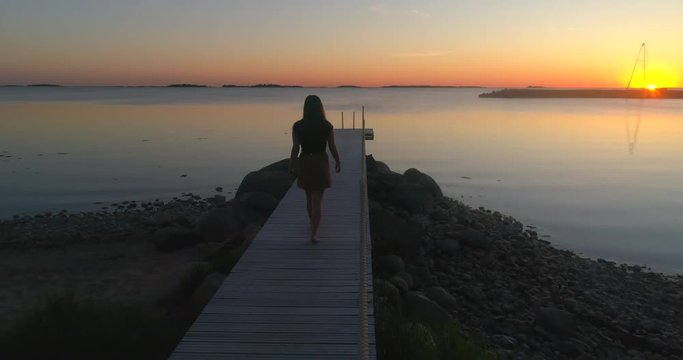 Woman on a wharf, C4K aerial tracking view a woman, walking from a wooden dock, enjoying the sun and a warm summer sunset, on a sunny evening dusk, in Hanko, Uusimaa, Finland