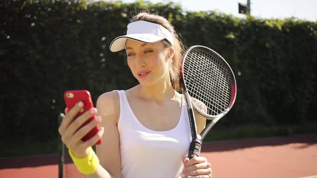 Beautiful cheerful young woman makes a video call while being on the tennis court, shows around, presents the tennis racket, waves to the screen. Modern technologies, gadgets, devices. Close up view