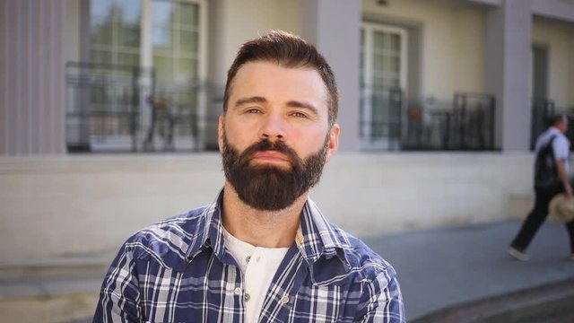 Brutal young bearded man in stylish checkered shirt raises his eyebrows in surprise, crosses his arms on his chest. Power, muscles, feeling confident. Outdoor shooting, sunny weather. Male portrait
