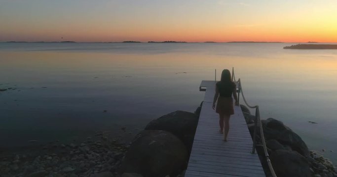 Woman on a wharf, C4K aerial view over a woman, standing at a wooden dock, enjoying the sun and a warm summer sunset, on a sunny evening dusk, in Hanko, Uusimaa, Finland
