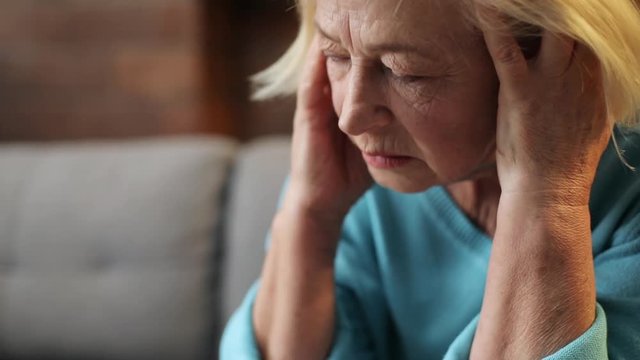 Cute unhappy Caucasian blonde-haired woman wearing blue sweater touching temples. Face of old upset woman. Close-up. Indoors. Illness.