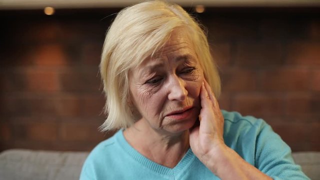Old sad Caucasian lady in blue sweater looking away, touching head. Portrait of lonely grandmother with toothache. Close-up. Indoors.