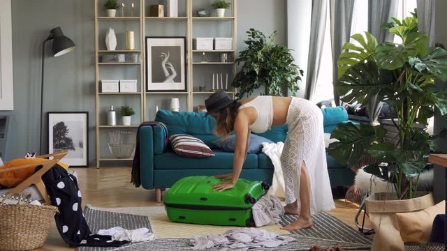 Beautiful woman in thirties of fair complexion packs suitcase for vacation with efforts and daydreams in stylish airy room with sophisticated interior and extra clothes scattered all around the place