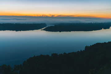 View from high shore on broad river. Riverbank with forest under thick fog. Dawn reflected in water. Yellow glow in picturesque predawn sky. Colorful morning atmospheric landscape of majestic nature.