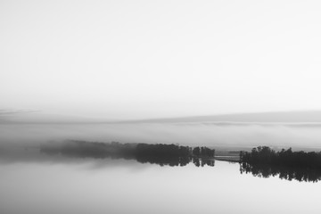 Fototapeta premium Broad mystical river flows along diagonal shore with silhouette of trees and thick fog in grayscale. Morning milky atmosphere. Minimalistic monochrome landscape of majestic nature.