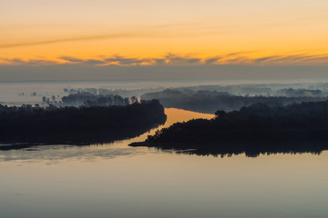 Riverbank with forest under predawn sky. Early blue sky reflected in river water. Yellow stripe in picturesque sky. Fog hid trees on island. Mystical morning atmospheric landscape of majestic nature.
