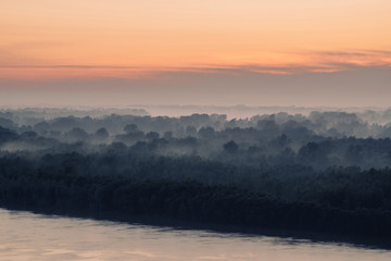 Mystical view on riverbank of large island with forest under haze at early morning. Eerie mist among layers from tree silhouettes. Morning atmospheric landscape of majestic nature in blue faded tones.