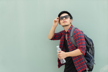 Portrait fashion handsome Asian man on grey background. Hipster traveler with backpack holding plastic water bottle and adjust sun glasses. Young male tourist hiker traveler on vacation.