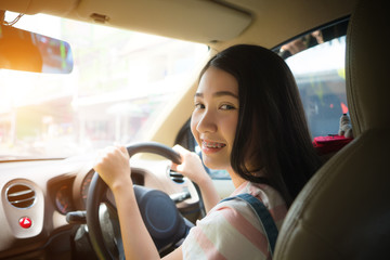 Young Asian woman driving car keeps wheel turning around smiling looking at passengers in back seat against sunset rays Light shine. Vehicle exam concept. Beautiful Smiling driver girl.