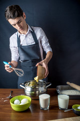 Young man chef in black apron cooking cake with ingredients on the table in the kitchen. Concept of men cook desserts