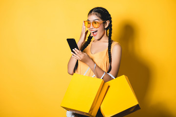 Beautiful young woman with shopping bags using her smart phone on yellow background.Shopaholic...