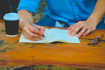 In the morning . Men sitting in the park  and he writing notebook. He drinking coffee and working on idea on notebook in relax time .