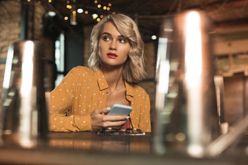 Inviting to bar. Pretty young woman using her phone and texting while sitting at the bar counter and occasionally looking at the entrance