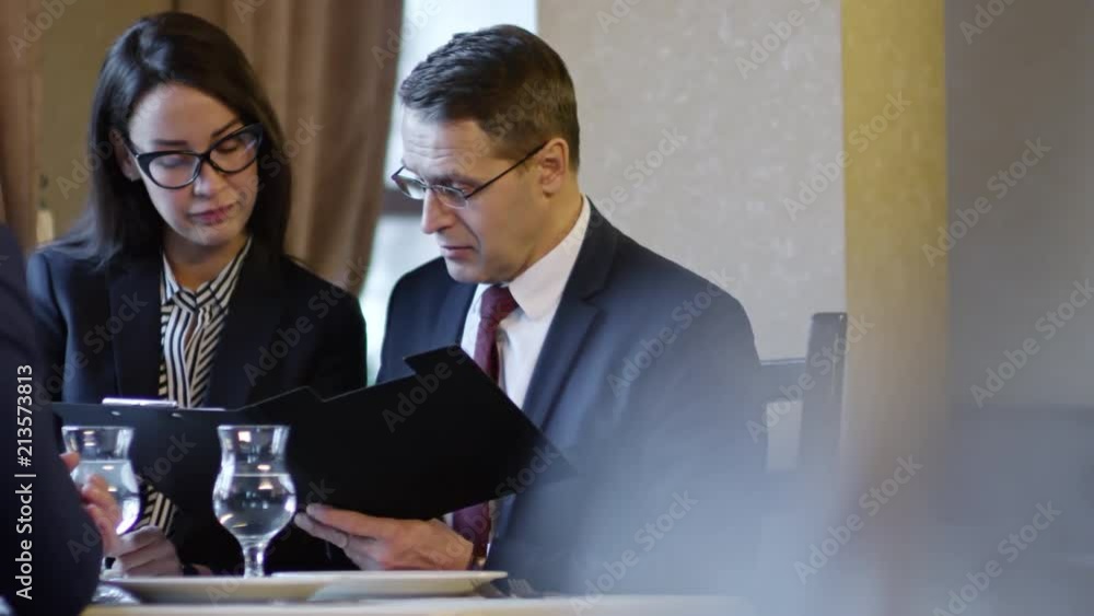 Poster tracking shot of three colleagues in business suits sitting at table in cafe or restaurant and discu