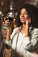 Lets meet. Gorgeous curly woman talking on the phone and inviting her friend to have a drink together while drinking wine at the bar