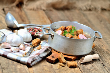 Old saucepan with garlic soup, fresh garlic bulbs, cloves, colored pepper, wooden spoon, croutons, lable and a towel on a rustic wooden table
