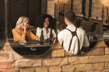 Obraz premium Pleasant interaction. Two beautiful female friends sitting at the bar counter and chatting with barman while drinking beer