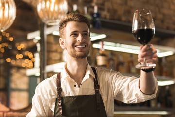 There you go. Upbeat young barman giving a glass of red wine to his customer and smiling at them brightly
