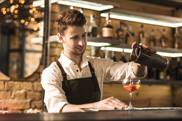 Skillful worker. Charming young barman standing at the bar counter and pouring a cocktail into the glass