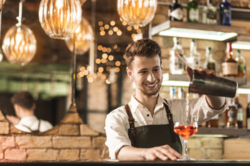 Nice cocktail. Handsome young barman standing behind the bar counter and filling the glass with a cocktail from shaker