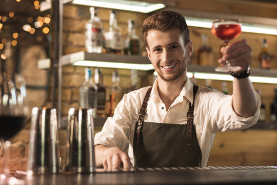 There you go. Pleasant young barman standing behind the bar counter and handing a cocktail to the customer while smiling