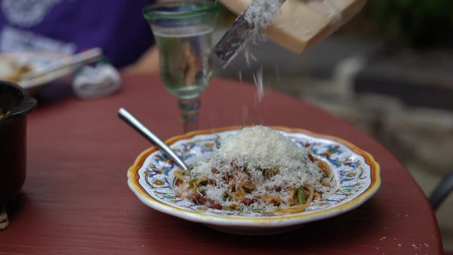 Cinemagraph of a cheese being grated onto an italian bowl of pasta with a glass of white wine in the distance.