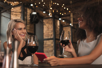 Amusing stories. Cheerful young women sitting at the bar counter, drinking wine and laughing heartily while sharing stories